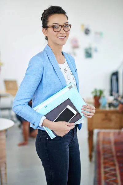 Shes the first one in and the last one out. Portrait of an attractive young woman carrying paperwork while standing in an office