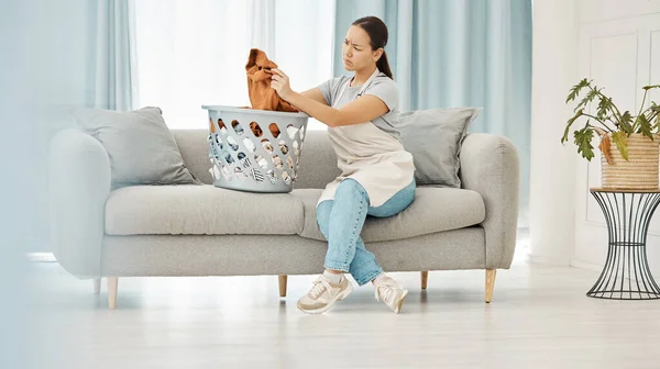 Asian woman, laundry or clothes basket on sofa in house living room and looking confused at dirty stains. Stress, anxiety or worry of person with washing clothing or cleaning fabric in home interior.