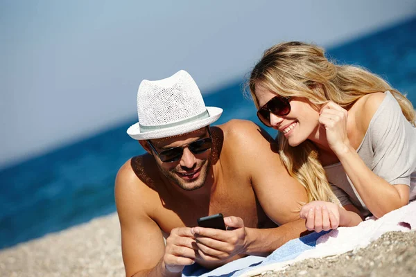 遠くにいるときは連絡を取り合っています ビーチで日焼けしている間に携帯電話を使っている若いカップルは — ストック写真