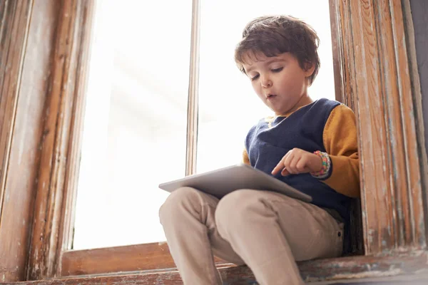 他的技术已经很熟练了 一个可爱的小男孩坐在窗台上用平板电脑 — 图库照片