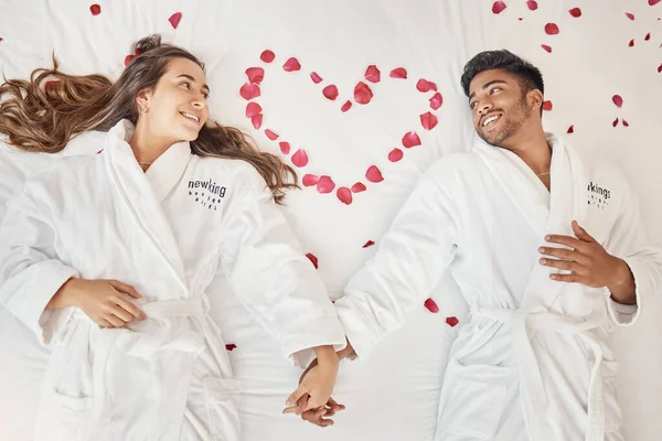 Man and woman married couple on luxury bed celebrate honeymoon, love and relax in romantic vacation spa hotel. Happy, marriage and young people smile on valentines day with rose heart in bedroom.