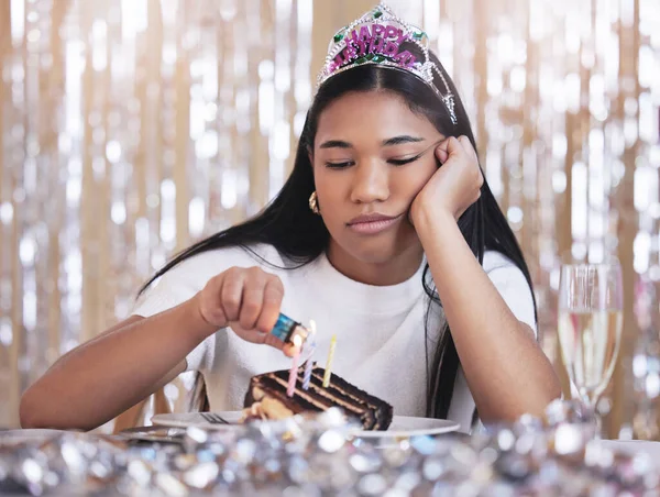 Sad, depression and angry woman at birthday celebration while lighting candle on cake. Lonely, depressed and frustrated female sitting at a party, fail and table with snack, decoration and tiara.