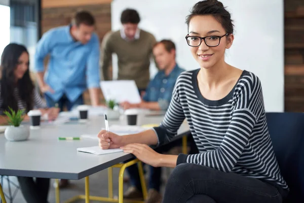 She stands out in the office. Portrait of an attractive young woman sitting in an office with colleagues in the background