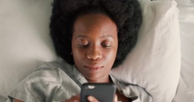 Telefonu olan bir kadın, sabah yatakta ve internette sosyal medyada, mesajlarda ve mesajlarda geziniyor. Siyah kadın akıllı telefona pijamalarıyla bakıyor ve yatak odasında yatakta uzanıyor..