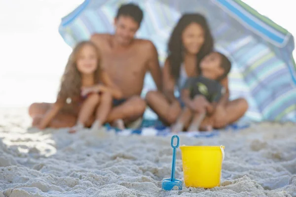 Having buckets of fun at the beach. A happy family smiling while sitting under an umbrella at the beach with a spade and bucket in the foreground