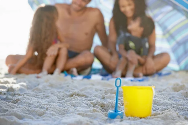 Spending quality time together. Cropped image of a family smiling while sitting under an umbrella at the beach with a spade and bucket in the foreground