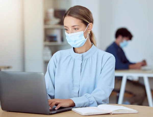 Laptop, face mask and corporate employee working on a project while sitting at a desk in the office. Professional woman typing company documents on her computer during a global covid pandemic