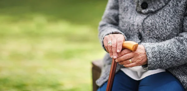 Old woman, hands and cane on park bench or nature in retirement. Senior retired disabled female pensioner, wood walking stick and wooden mobility aid support for balance and old age problem