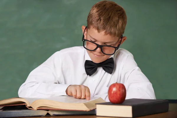 彼は読むのが好きだ 彼の前のテーブルに赤いリンゴと彼の読書に集中眼鏡と蝶ネクタイを身に着けている若い男の子 — ストック写真