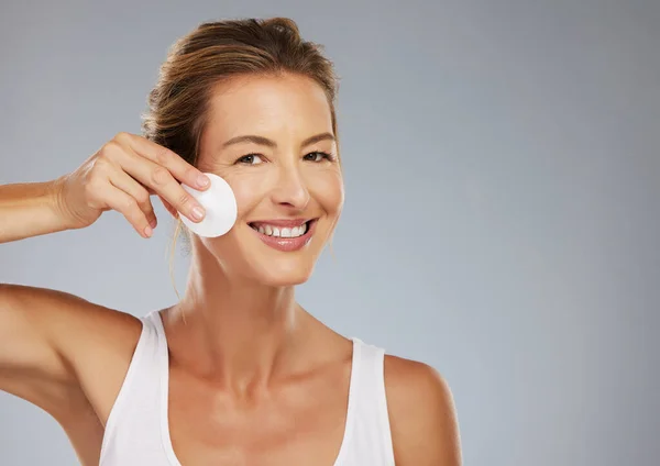 Skincare, mature woman and facial cotton cleaning face, makeup or cosmetics removal on a gray studio background. Wellness, health and female model with skin cleanser, beauty treatment or exfoliation.