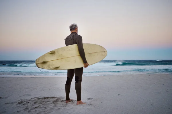 Surfer, surfboard and senior man on beach at sea waves in during sunset during summer vacation in Hawaii. Professional male athlete rest after training or practice surfing sport outdoor at the ocean.