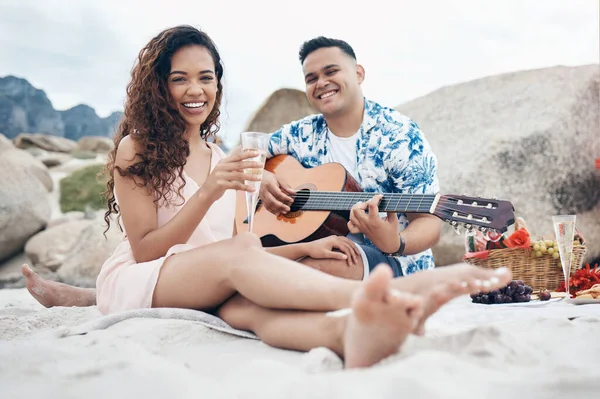 Couple, beach and guitar for music and love on picnic together on sand by ocean. Man, woman and smile with happiness on face with food, champagne and flowers to relax, vacation and travel in summer.