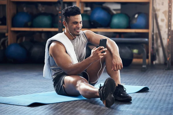Man, exercise and phone on social media with smile rest on floor in gym during training session. Guy, wellness and smartphone take break during workout for fitness, health and sport with happiness.