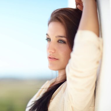 Gazing into the distance. A beautiful young woman leaning on a white wall outside clipart