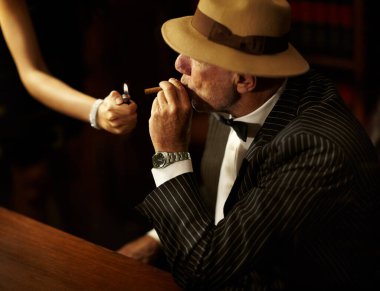 Başkaları üzerinde gücü ve etkisi var. Bir kadın onun için sigara yakarken, yaşlı mafya babası bir şapka takıyor ve ciddi görünüyor.
