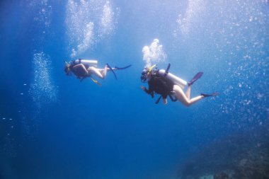 Derin maviliğe dalmış. İki skuba dalgıcı etraflarındaki kabarcıklarla birlikte derin mavi okyanusta huzur içinde yüzerler.