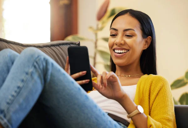西班牙裔女人一边笑着一边用智能手机在明亮的客厅里放松 一名年轻女子在家中躺在沙发上 用现代技术在手机上打滚 — 图库照片