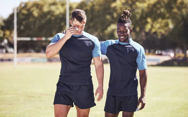 Two diverse young rugby players standing outside on the field. Black man smiling while talking to his injured and bruised caucasian teammate. Athletic sportsmen discussing and reflecting on the game.