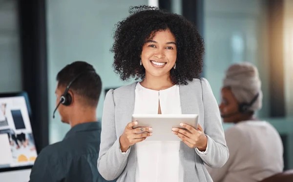 Portrait of young mixed race businesswoman browsing on a digital tablet device while working in a call centre with her colleagues in the background. Happy manager and supervisor planning online with .