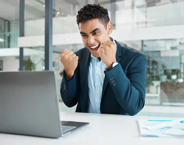 Young mixed race businessman cheering with passion while working on a laptop alone at work. One hispanic businessperson smiling and celebrating victory working at a desk in an office.