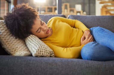 Young unhappy mixed race woman holding her stomach and suffering from stomach pain on the couch at home. One sick hispanic woman suffering from cramps resting on the sofa. clipart