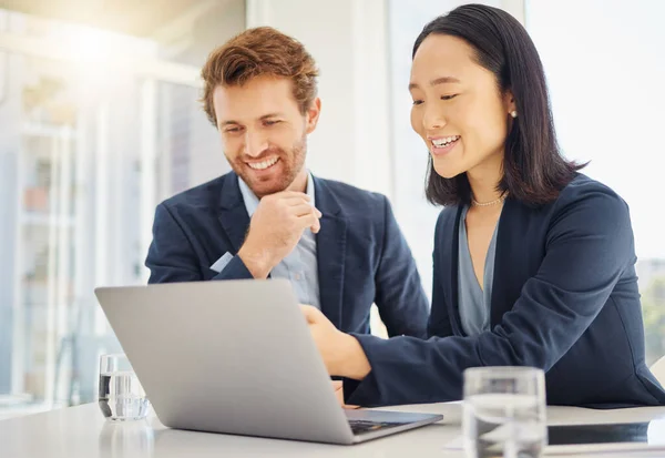 Two happy young diverse colleagues working together on a laptop in an office boardroom. Confident asian businesswoman explaining corporate plans and strategy to smiling caucasian businessman during a.