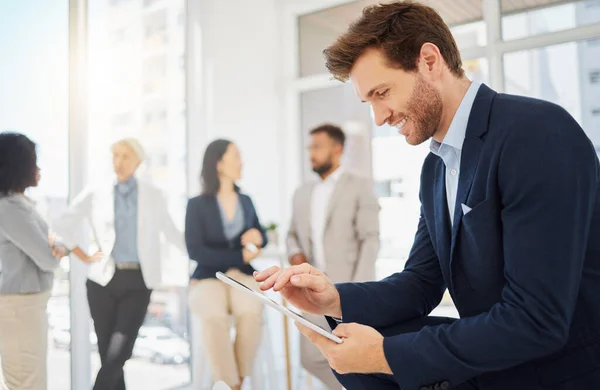 Young caucasian businessman browsing on a digital tablet device in an office with his diverse colleagues in the background. Man searching online for ideas and using smart apps to prepare for his next.