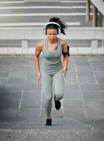 Young mixed race female wearing gym wear and wireless headphones while running up the steps of a building outside. Young female focused on her speed, body, fitness and cardio health while training.