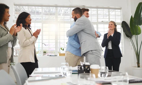 Happy businessmen hugging each other in support and unity while colleagues clap hands in a meeting at work. Diverse group of cheerful businesspeople welcoming an employee to their workplace together.