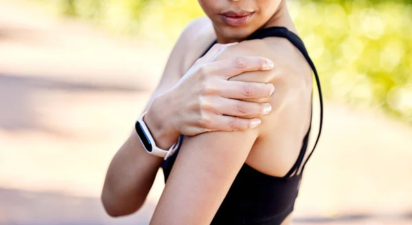 Closeup of one mixed race woman holding her sore shoulder while exercising outdoors. Female athlete suffering with painful arm injury from fractured joint and inflamed muscles during workout. Struggl.