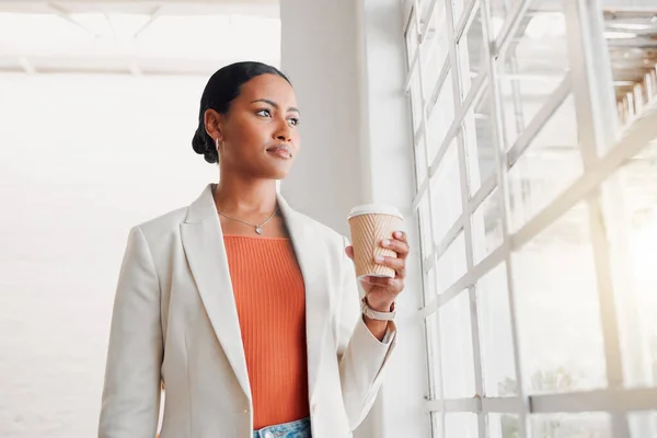 Young mixed race businesswoman drinking a coffee alone in an office at work. Confident hispanic businessperson looking out of a window drinking a coffee standing at work.