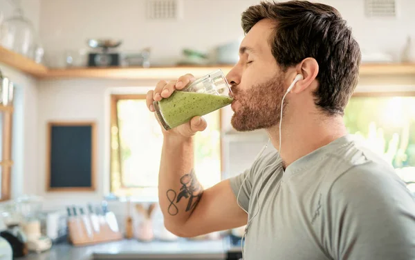 One fit young caucasian man drinking a glass of healthy green detox smoothie while wearing earphones in a kitchen at home. Guy having fresh fruit juice to cleanse and provide energy for training. Who.