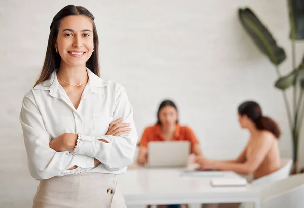 Portrait of one confident young hispanic business woman standing with arms crossed in an office with her colleagues in the background. Ambitious entrepreneur and determined leader ready for success i.