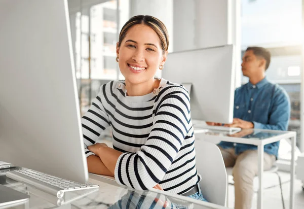 Portrait of a young mixed race businesswoman using a desktop computer in an office at work. Happy hispanic female businessperson smiling while sitting at a desk. Business professional working on a co.