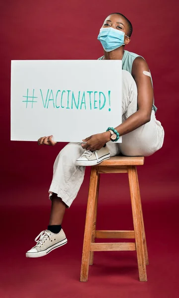 アフリカ系アメリカ人の流行の予防接種を受けた女性が腕の石膏を見せ ポスターを持ち 外科用フェイスマスクを着用した コピースペース付きの赤いスタジオの背景に隔離された幸せな黒いモデル コロナワクチンはSigを促進する — ストック写真