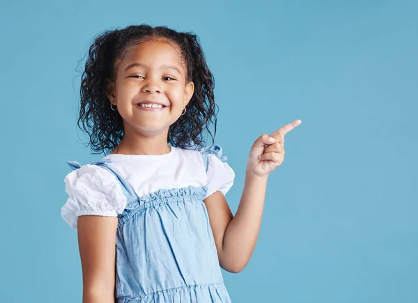 Portrait of happy smiling little girl pointing her finger at copy space to the right against blue studio background. Cheerful mixed race kid in casual clothes.