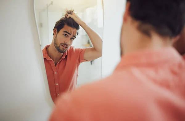 Handsome young caucasian man touching his hair and looking in the bathroom mirror. Male pulling his hair and thinking of getting a haircut. Concerned man worried about dandruff, receding hairline or .