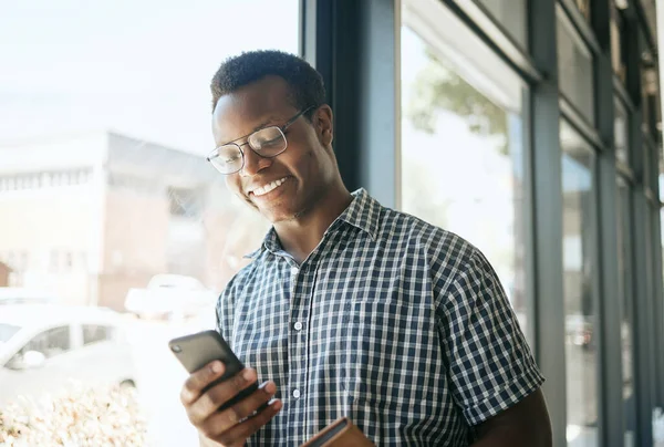Happy african american business man entrepreneur leaning against a window in an office while reading or sending text on smartphone. Smiling entrepreneur chatting online or getting good news via app.