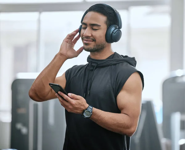 Smiling asian trainer alone in gym, listening to music on headphones from cellphone. Handsome coach standing in health club workout, selecting song from playlist on technology. Man in fitness centre.