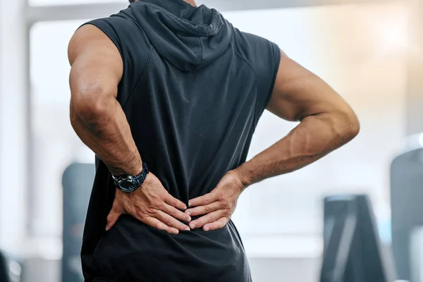 Unknown trainer alone in gym and suffering from back injury. Coach standing and rubbing back during workout in exercise health club. One man in fitness centre feeling backache pain in routine trainin.