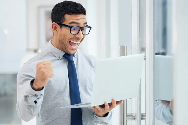 Amazed young mixed race businessman standing and working on a laptop in an office at work. Joyful hispanic male businessperson looking shocked while cheering and holding a laptop at work.