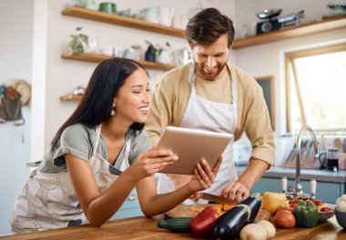 Evde birlikte yemek pişirirken dijital tablet kullanan mutlu, genç bir çift. İspanyol karısı internette yemek tarifi okurken, genç beyaz bir adam sebze kesiyor..