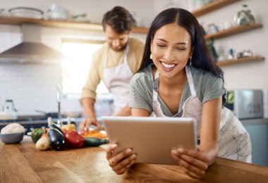 Erkek arkadaşı arka planda yemek pişirirken, genç mutlu kadının dijital tablete göz atmasını kapat. Kocasıyla evde yemek pişirirken internetten yemek tarifi arayan genç İspanyol kadın..