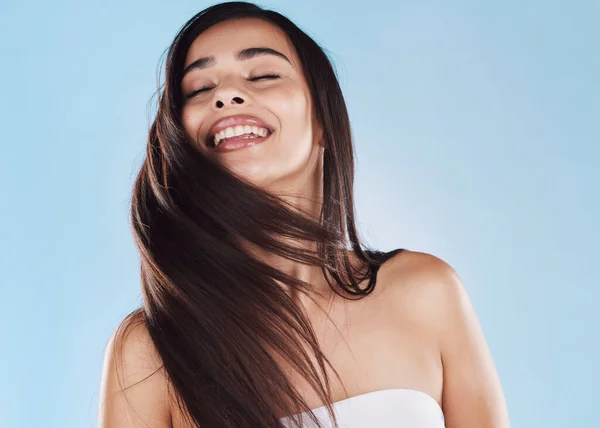 One beautiful young hispanic woman with healthy skin tossing her sleek long hair against a blue studio background. Happy mixed race model with flawless complexion and natural beauty.