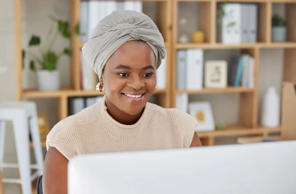 Young african american businesswoman with a headscarf sitting alone in an office and browsing the internet on a computer. Ambitious black creative professional networking and emailing clients at desk.