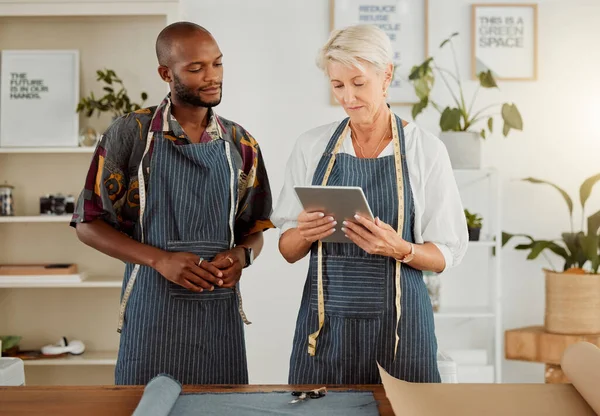 Two designers using a digital tablet while working with material at work. Mature caucasian tailor talking and holding a digital tablet while working with a male african american colleague.
