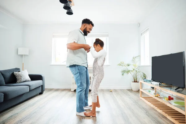 A mixed race father dancing with his daughter while she stands on his feet in the lounge at home. Hispanic male having fun with his daughter in the living room.