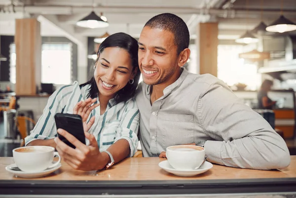 Happy young mixed race couple sitting at table having coffee while looking at something on smartphone in cafe. Loving couple smiling while taking selfie or doing video call on mobile phone while on a.