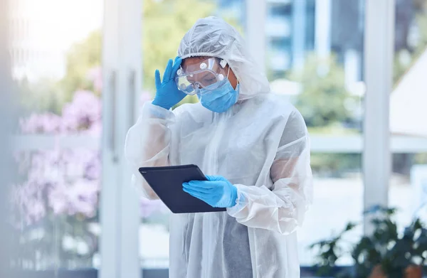 Stressed and worried woman using digital tablet while wearing a protective hazmat suit to keep safe from covid and disease in hospital or lab. One female only struggling with a headache and challenge.