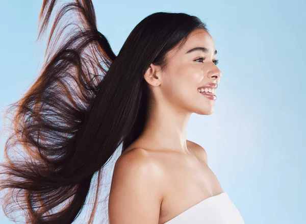 One beautiful young hispanic woman with healthy skin and sleek long hair blowing in the wind smiling against a blue studio background. Happy mixed race supermodel with flawless complexion and natural.